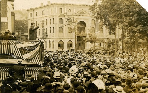 A large crowd gathers outdoors, listening to a speaker on a platform, with historic buildings in the background.