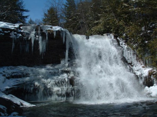 A frozen waterfall surrounded by snow-covered rocks and trees, with icicles hanging from the ledge.