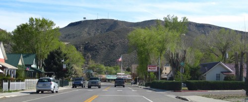 A small town street lined with trees, leading to a hill with a radio tower in the background under a clear blue sky.