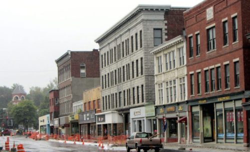 A street view of historic buildings with shops, under overcast skies and construction barriers in the foreground.