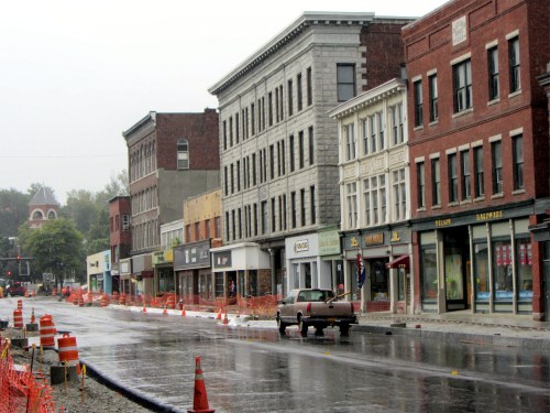 A rainy street scene with historic buildings, construction barriers, and a parked car on a wet road.
