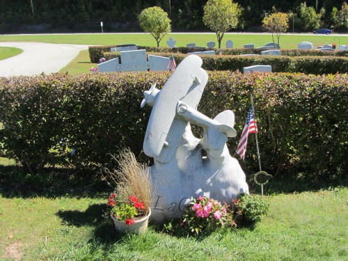 A unique gravestone shaped like an airplane, surrounded by flowers and an American flag in a cemetery.
