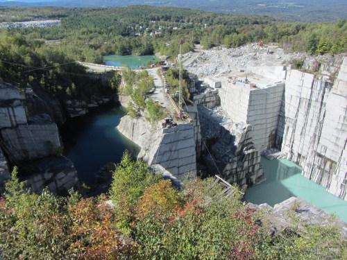 Aerial view of a granite quarry with turquoise water, surrounded by trees and rocky cliffs under a clear blue sky.