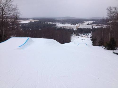 A snowy landscape with ski slopes and trees, overlooking a valley and distant hills under a cloudy sky.