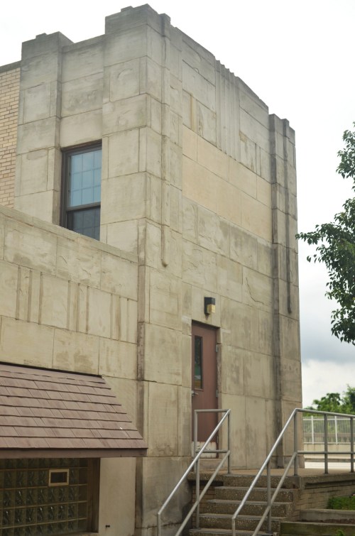 A close-up of a textured concrete building corner with a door and stairs, surrounded by greenery.