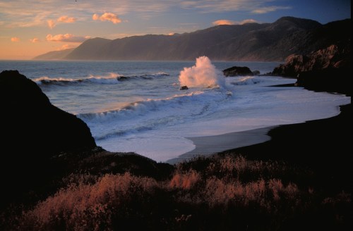 A serene coastal scene at sunset, with waves crashing on a rocky shore and mountains in the background.
