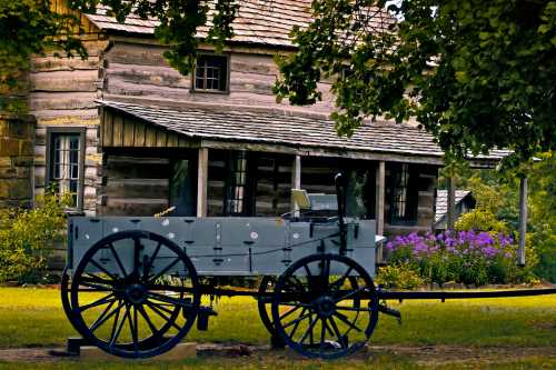 A vintage wagon in front of a rustic wooden house, surrounded by greenery and blooming flowers.