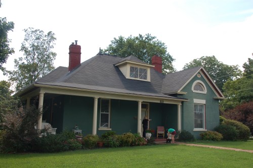 A green house with a porch, surrounded by trees and a lawn, featuring a gabled roof and red chimneys.