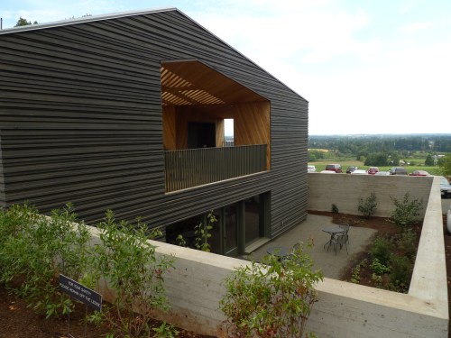 Modern two-story house with a wooden balcony, surrounded by greenery and a concrete wall, overlooking a scenic landscape.