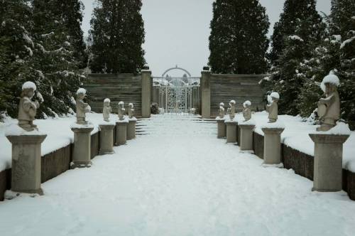 A snowy pathway lined with statues leads to a gated entrance, surrounded by tall evergreen trees.