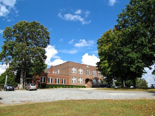 A brick building surrounded by trees and a gravel parking area under a blue sky with scattered clouds.