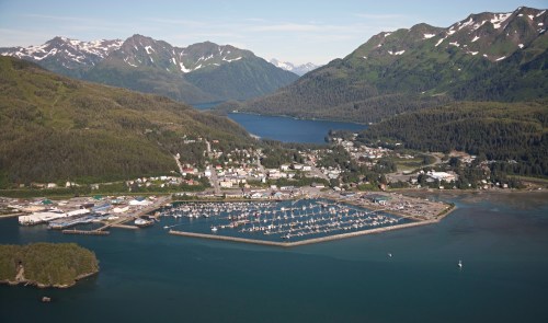 Aerial view of a coastal town surrounded by mountains, featuring a marina filled with boats and lush greenery.