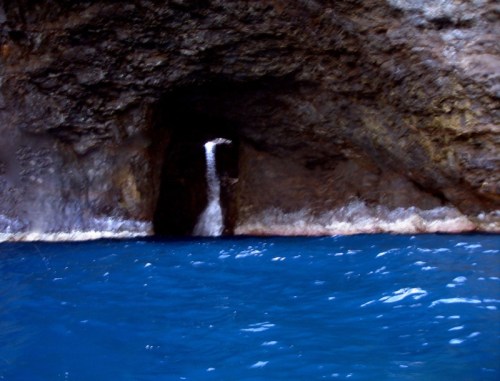 A rocky cave opening with a small waterfall, surrounded by deep blue water.