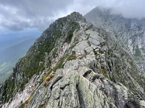 A rocky mountain ridge under a cloudy sky, with lush green valleys visible in the distance.
