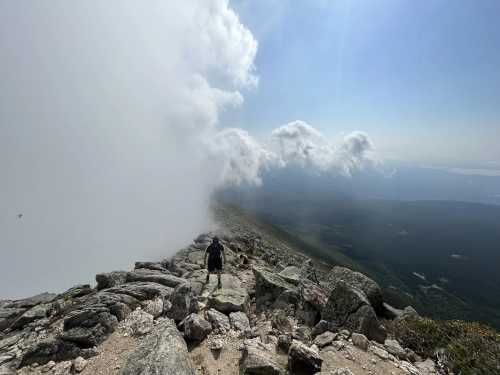 A hiker navigates rocky terrain on a mountain, with clouds rolling in and a vast landscape visible below.