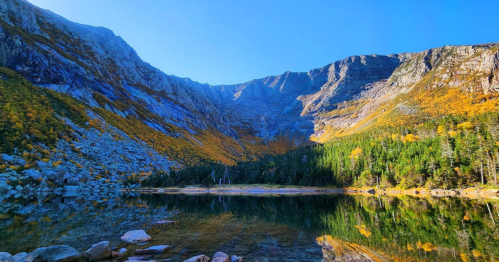 A serene mountain landscape with a clear lake reflecting vibrant autumn foliage and rocky cliffs under a blue sky.