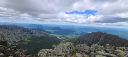 A panoramic view from a mountain peak, showcasing lush valleys, distant mountains, and a cloudy sky.