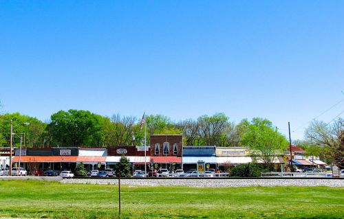 A sunny day view of a small town street lined with shops and greenery in the background.