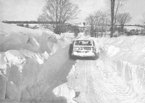 A car navigating a narrow path through high snowdrifts on either side, with trees in the background.