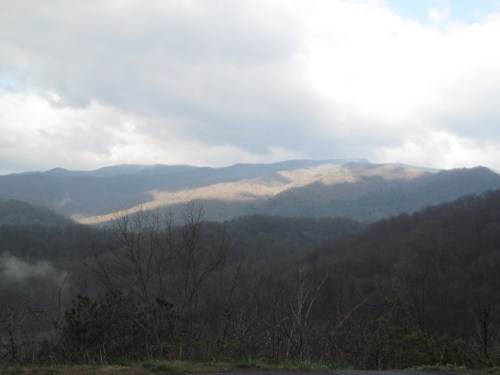 A scenic view of misty mountains under a cloudy sky, with sunlight illuminating the peaks in the distance.