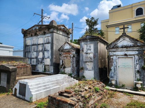 A historic cemetery with weathered tombs and mausoleums under a blue sky, surrounded by greenery and urban buildings.
