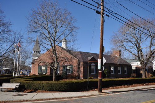 A brick building with a steeple, surrounded by trimmed hedges and trees, under a clear blue sky.