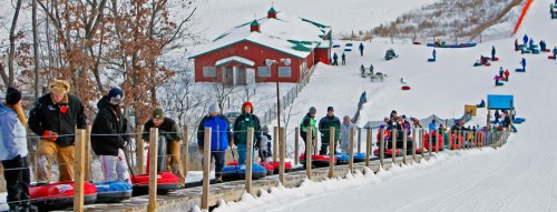 A snowy hill with people sledding and a conveyor belt transporting sleds, surrounded by trees and a red building.