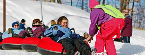 A group of people in winter gear prepares to go tubing down a snowy hill, assisted by an employee in bright clothing.