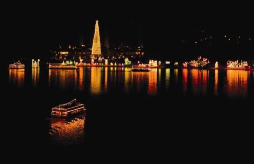 A festive scene of illuminated boats and a large Christmas tree reflecting on dark water at night.