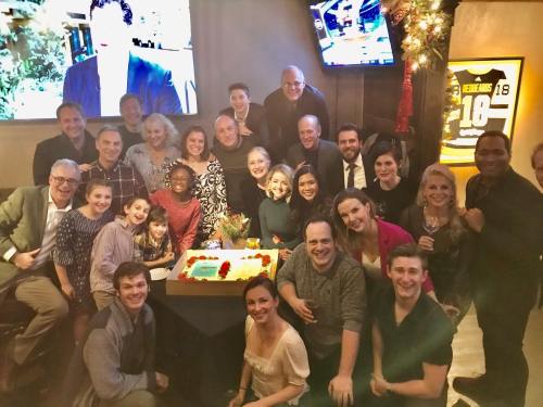 A large group of people gathered around a decorated cake, smiling and celebrating in a festive setting.