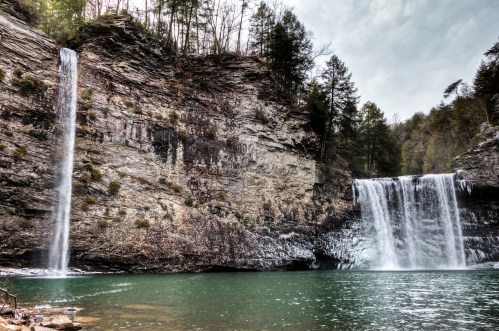 Two waterfalls cascade down rocky cliffs into a serene green pool, surrounded by lush trees and a cloudy sky.