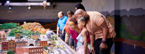 A family observes a detailed miniature train display, admiring the intricate scenery and models.