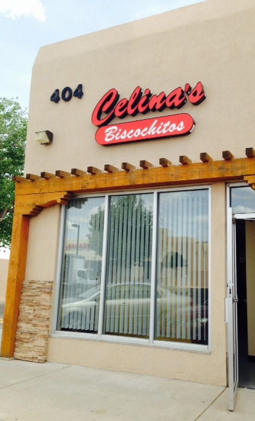 Exterior of Celina's Biscochitos shop, featuring a sign with the name and address, and large windows with vertical blinds.