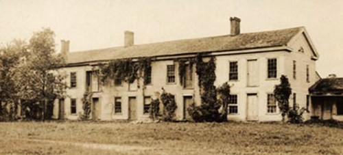 Historic two-story building covered in vines, set in a grassy area with trees nearby.