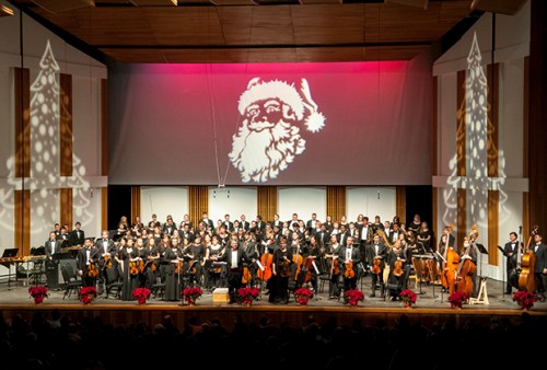 A festive orchestra performance on stage, decorated with Christmas trees and a Santa projection in the background.
