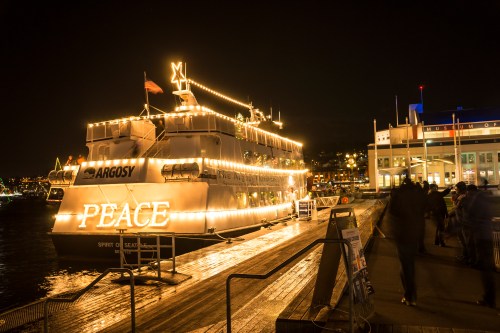 A brightly lit boat named "Peace" docked at night, with people walking along the pier nearby.