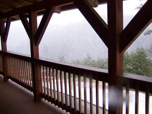 A snowy scene viewed from a wooden porch, with snowflakes falling and a blurred landscape in the background.