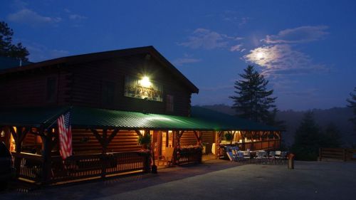 A log cabin at dusk, illuminated by moonlight, with an American flag and outdoor seating visible.