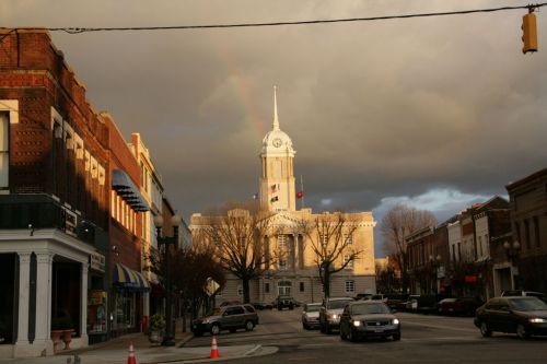 A historic courthouse stands at the end of a street, illuminated by sunlight against a backdrop of dark clouds and a rainbow.