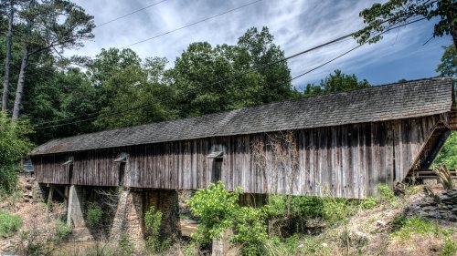A weathered wooden covered bridge spans a small creek, surrounded by lush greenery and trees under a cloudy sky.