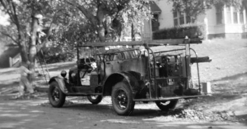 A vintage fire truck parked on a street, surrounded by trees and a house in the background.