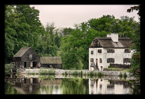 A serene landscape featuring a black wooden mill and a white house by a calm river, surrounded by lush greenery.