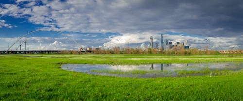 A panoramic view of a city skyline with a bridge, green fields, and a reflective pond under a cloudy sky.
