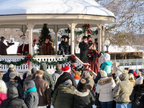 A festive crowd gathers around a decorated gazebo with performers in winter attire during a snowy holiday event.