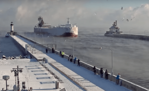 A snowy pier with people watching a large ship passing through a harbor, surrounded by seagulls and icy waters.