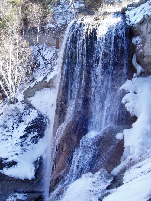 A frozen waterfall cascading down rocky cliffs, surrounded by snow-covered trees and a winter landscape.