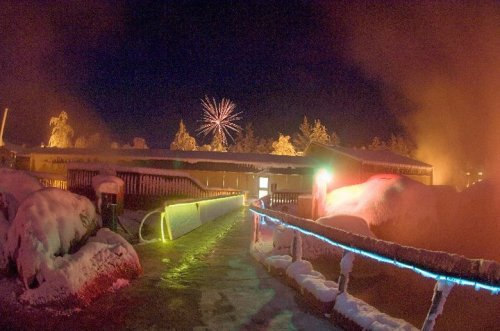 A snowy pathway leads to a building, illuminated by colorful lights, with fireworks in the night sky above.