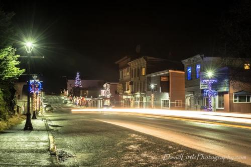 A quiet street at night, lined with festive lights and decorations, featuring a Christmas tree in the distance.