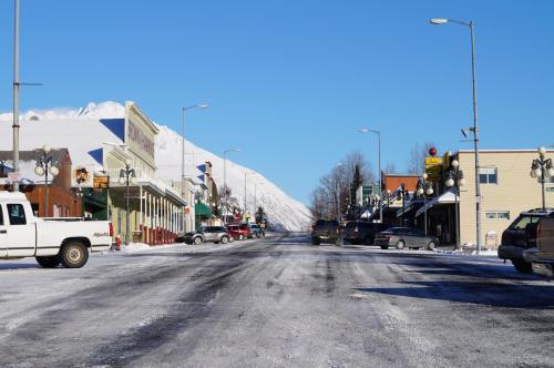 Snow-covered street lined with buildings and parked cars, under a clear blue sky and mountains in the background.