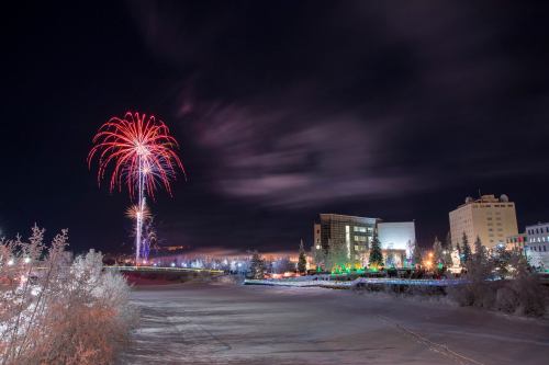 Colorful fireworks illuminate a night sky over a snowy landscape and buildings, creating a festive atmosphere.